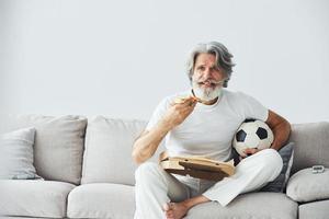 Soccer fan with pizza watches match. Senior stylish modern man with grey hair and beard indoors photo
