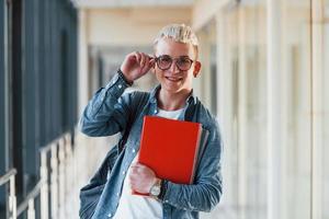 Male young student in jeans clothes is in corridor of a college with notepad in hands photo