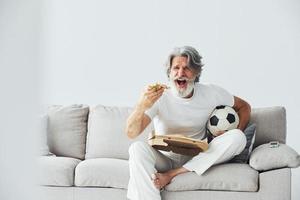 Soccer fan with pizza watches match. Senior stylish modern man with grey hair and beard indoors photo