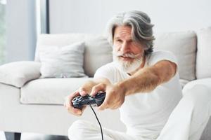 juega videojuegos usando el controlador. Senior hombre moderno con estilo con pelo gris y barba en el interior foto