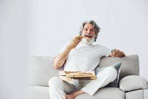 With delicious pizza. Watches TV show. Senior stylish modern man with grey hair and beard indoors photo