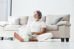 en auriculares. se sienta cerca del sofá. Senior hombre moderno con estilo con pelo gris y barba en el interior foto