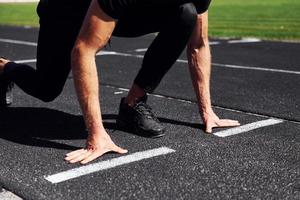 Runner is on starting position on track. Sportive young guy in black shirt and pants outdoors at daytime photo