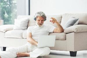 en auriculares. se sienta cerca del sofá. Senior hombre moderno con estilo con pelo gris y barba en el interior foto