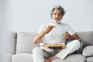 With delicious pizza. Watches TV show. Senior stylish modern man with grey hair and beard indoors photo