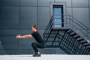 Exercising near building with black wall. Sportive young guy in black shirt and pants outdoors at daytime photo