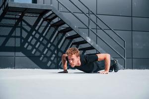 Doing push ups. Sportive young guy in black shirt and pants outdoors at daytime photo