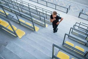 Running on the bleachers. Sportive young guy in black shirt and pants outdoors at daytime photo