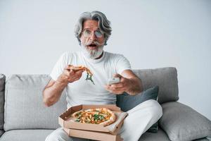 Eats delicious pizza while watching TV show. Senior stylish modern man with grey hair and beard indoors photo