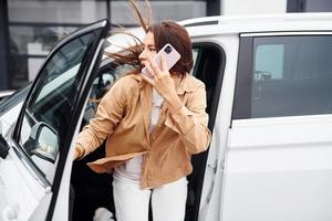 Talks by phone with door opened. Fashionable beautiful young woman and her modern automobile photo