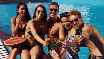 sosteniendo sandía. un grupo de jóvenes felices se divierten en la piscina durante el día foto