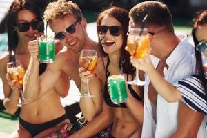 Fresh cocktails in hands. Group of young happy people have fun in swimming pool at daytime photo