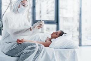 Female doctor in defensive lab coat and protective eyewear with syringe in hand injecting medicine to young girl sick of virus photo