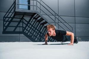 Doing push ups. Sportive young guy in black shirt and pants outdoors at daytime photo