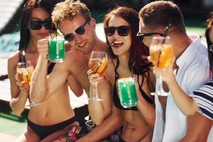 Fresh cocktails in hands. Group of young happy people have fun in swimming pool at daytime photo