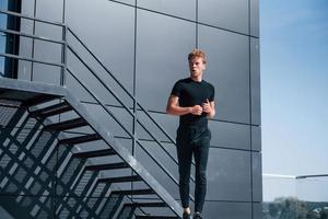 Warming up near black building. Sportive young guy in black shirt and pants outdoors photo