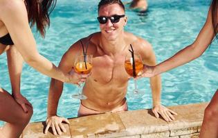 Fresh cocktails in hands. Group of young happy people have fun in swimming pool at daytime photo