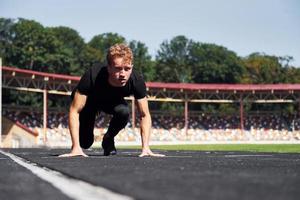 Runner is on starting position on track. Sportive young guy in black shirt and pants outdoors at daytime photo