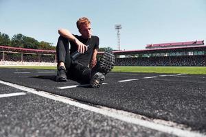 Tired runner sits on track and taking a break. Sportive young guy in black shirt and pants outdoors at daytime photo