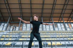 Jumping and doing parkour on the bleachers. Sportive young guy in black shirt and pants outdoors at daytime photo