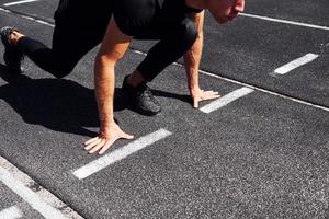 Runner is on starting position on track. Sportive young guy in black shirt and pants outdoors at daytime photo