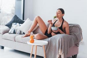 Young woman with slim body shape in sportswear sits on sofa and eats healthy diet food indoors at home photo