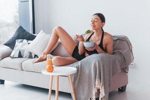 Young woman with slim body shape in sportswear sits on sofa and eats healthy diet food indoors at home photo