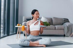 With healthy food and orange juice in hands. Young woman with slim body shape in sportswear have fitness day indoors at home photo