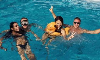 Using yellow colored duck to swim. Group of young happy people have fun in swimming pool at daytime photo