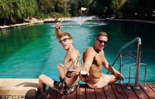 Two male friends sits near water outdoors with drinks in hands photo