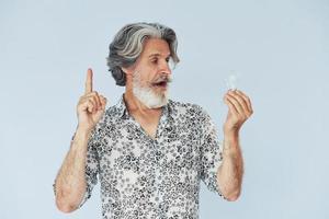 Holds light bulb in hands. Conception of ideas and inspiration. Senior stylish modern man with grey hair and beard indoors photo