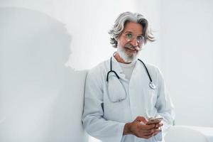 Senior male doctor with grey hair and beard in white coat is indoors in clinic photo