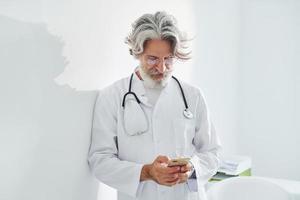 Senior male doctor with grey hair and beard in white coat is indoors in clinic photo