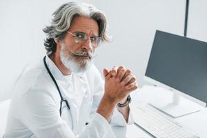 retrato de un médico de alto rango con cabello gris y barba en bata blanca está adentro en la clínica foto