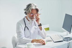 Writes by computer table. Senior male doctor with grey hair and beard in white coat is indoors in clinic photo