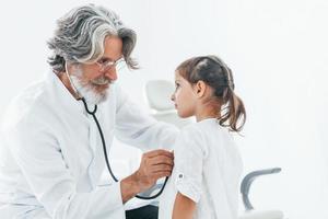 Senior male doctor with grey hair and beard in white coat measuring heart rate of little girl by stethoscope indoors in clinic photo