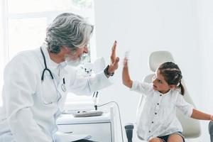 Working with little girl. Giving high five. Senior male doctor with grey hair and beard in white coat is indoors in clinic photo