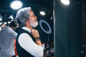 en ropa retro en barbería. un anciano moderno y elegante con pelo gris y barba está en el interior foto