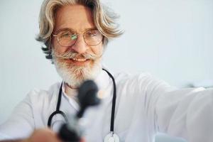 retrato de un médico de alto rango con cabello gris y barba en bata blanca está adentro en una clínica de otorrinolaringología foto