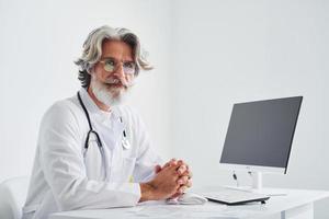 Portrait of senior male doctor with grey hair and beard in white coat is indoors in clinic photo