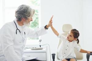 Working with little girl. Giving high five. Senior male doctor with grey hair and beard in white coat is indoors in clinic photo