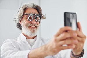 un anciano con cabello gris y barba está en una clínica de oftalmología sonríe y sostiene un teléfono inteligente foto