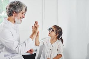 Oftalmólogo de sexo masculino senior con cabello gris y barba en bata blanca está adentro en la clínica probando la vista de la niña foto