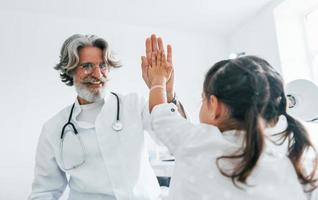 Working with little girl. Giving high five. Senior male doctor with grey hair and beard in white coat is indoors in clinic photo