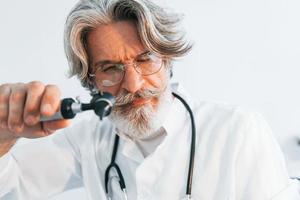 Portrait of senior male doctor with grey hair and beard in white coat is indoors in otorhinolaryngology clinic photo