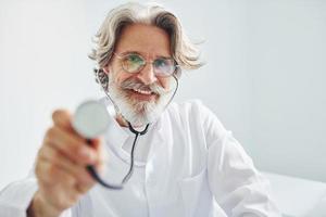 Holds stethoscope. Portrait of senior male doctor with grey hair and beard in white coat is indoors in clinic photo