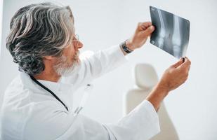 Looks at x-ray. Senior male doctor with grey hair and beard in white coat is indoors in clinic photo