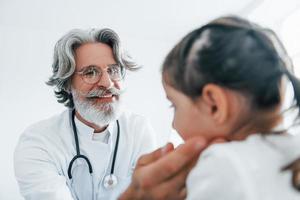 Little girl have a visit. Senior otorhinolaryngologist with grey hair and beard in white coat is indoors in clinic photo