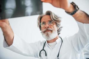 Looks at x-ray. Senior male doctor with grey hair and beard in white coat is indoors in clinic photo