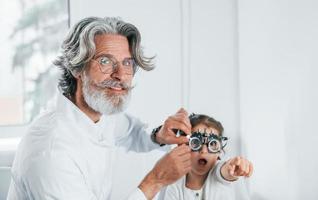 Senior male ophthalmologist with grey hair and beard in white coat is indoors in clinic testing sight of little girl photo